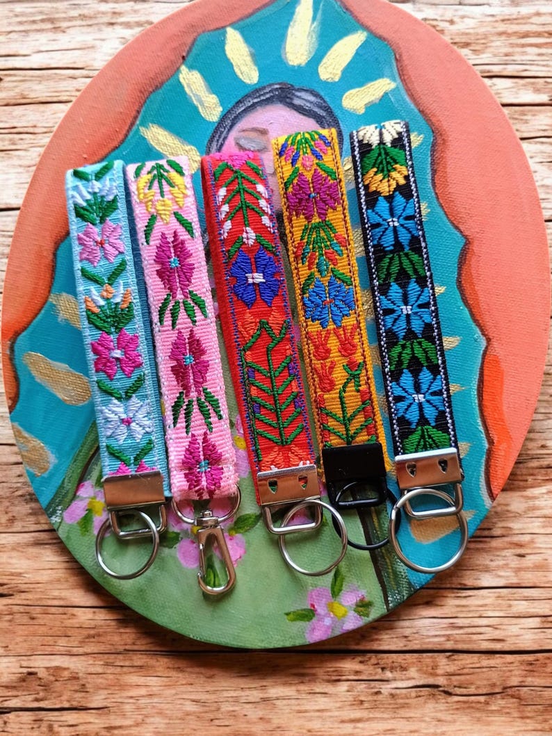 May include: Five fabric wristlet keychains featuring intricate floral embroidery in light blue, pink, red, yellow, and black. Each strap is finished with a silver-toned metal clasp and a circular split ring, except for the yellow one with a black clasp.