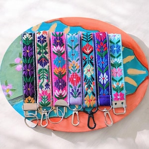 May include: Seven fabric wristlet keychains featuring vibrant, multi-colored floral embroidery patterns. Each strap is finished with a metal clasp, including silver-toned rings, lobster claw clips, and one black clip, attached to a decorative base.