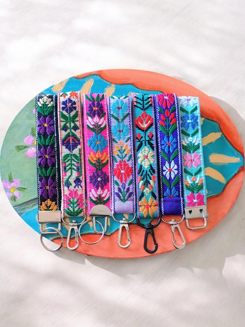 May include: Seven fabric wristlet keychains featuring vibrant, multi-colored floral embroidery patterns. Each strap is finished with a metal clasp, including silver-toned rings, lobster claw clips, and one black clip, attached to a decorative base.