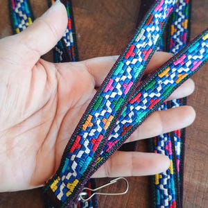 May include: A long, narrow fabric strap featuring a vibrant, multi-colored woven pattern with zig-zag lines in shades of blue, green, red, yellow, and white against a dark brown base, finished with a silver-toned metal swivel hook clasp.