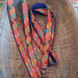 May include: A long, narrow fabric strap featuring a vibrant, multi-colored woven pattern in shades of red, orange, green, and blue. The strap is finished with a dark blue fabric backing and includes silver-toned metal swivel clips at the ends.