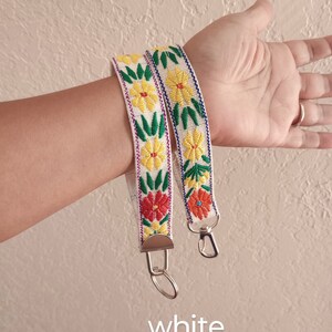 May include: A white fabric wristlet strap featuring embroidered yellow and red flowers with green leaves, finished with silver-toned metal hardware including a lobster clasp and a split ring. The word white is printed at the bottom of the image.