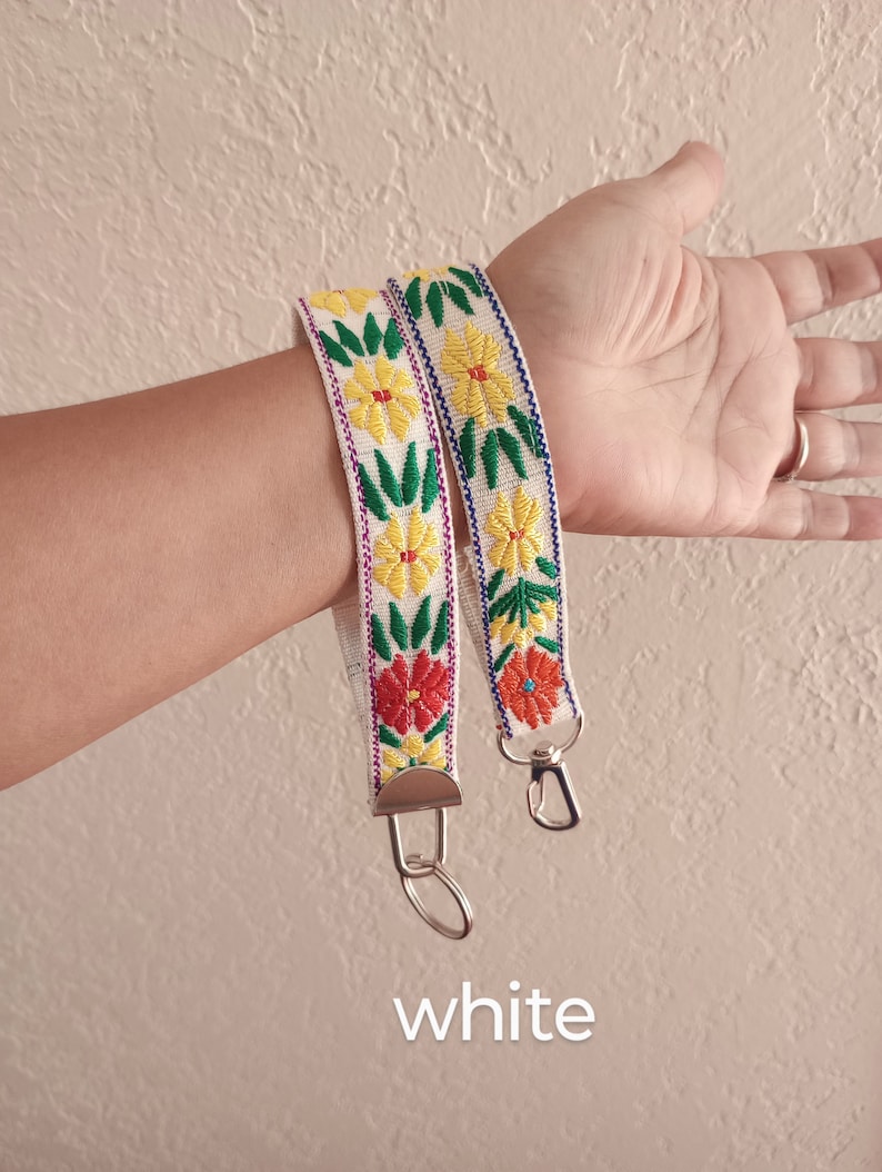 May include: A white fabric wristlet strap featuring embroidered yellow and red flowers with green leaves, finished with silver-toned metal hardware including a lobster clasp and a split ring. The word white is printed at the bottom of the image.