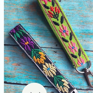 May include: Two fabric wristlet keychains with embroidered floral patterns. One keychain is moss green with pink and red flowers, and the other is black with purple, yellow, and green flowers. Both feature silver metal hardware and key rings.