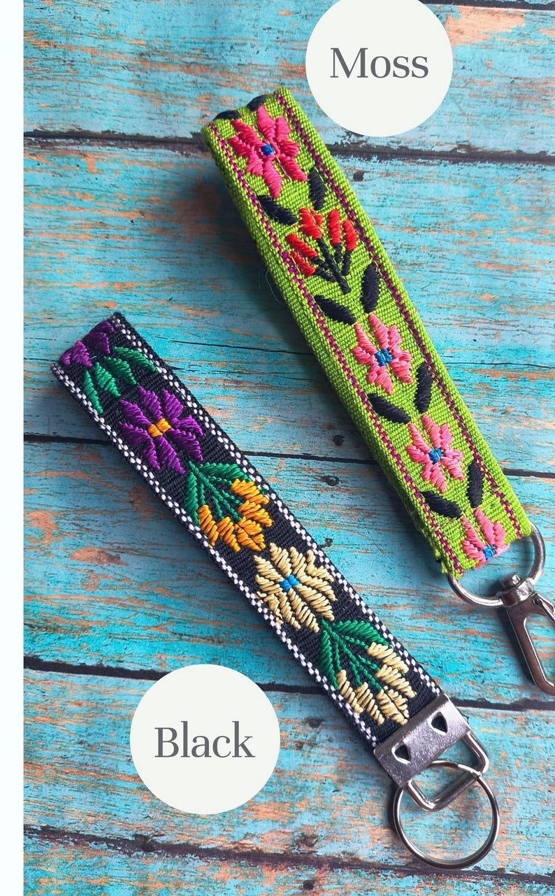May include: Two fabric wristlet keychains with embroidered floral patterns. One keychain is moss green with pink and red flowers, and the other is black with purple, yellow, and green flowers. Both feature silver metal hardware and key rings.