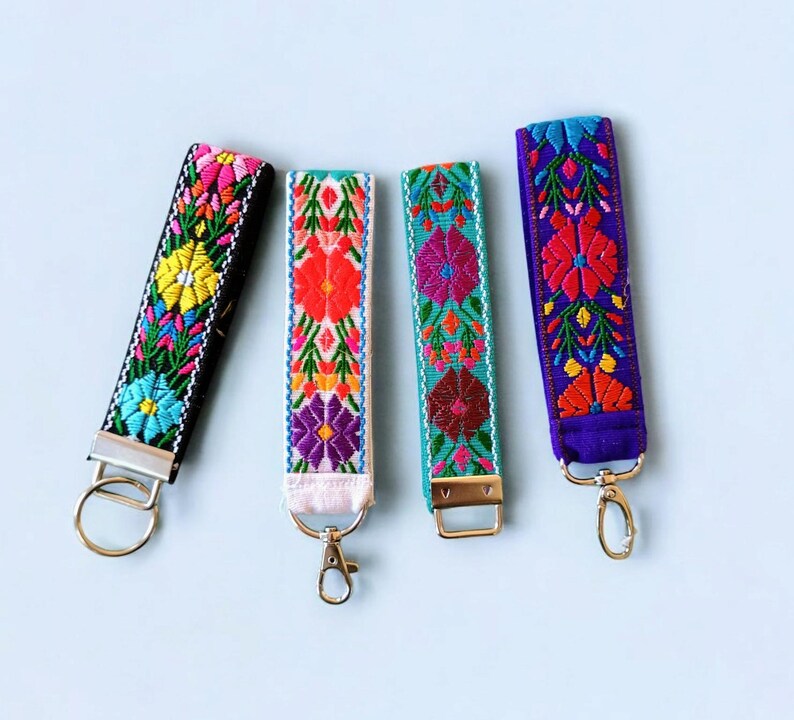 May include: Four colorful embroidered keychains with floral patterns. The keychains are black, white, turquoise, and purple. Each keychain has a silver key ring.