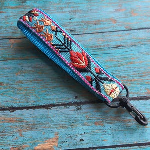 May include: A turquoise fabric wrist strap with embroidered floral designs in red, orange, and white. The strap has a black clasp and is approximately 1 inch wide. The background is a distressed blue wooden surface.