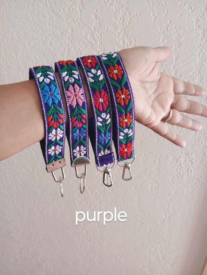 May include: Four purple fabric wristlet keychains featuring a woven floral pattern in shades of red, pink, blue, green, and white. Each strap is finished with a silver-toned metal clasp and a circular split ring for attaching keys or accessories.