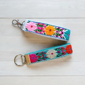 May include: Two colorful embroidered keychains with metal rings. One keychain is white with pink, orange, and yellow flowers. The other keychain is turquoise with pink, orange, and white flowers.
