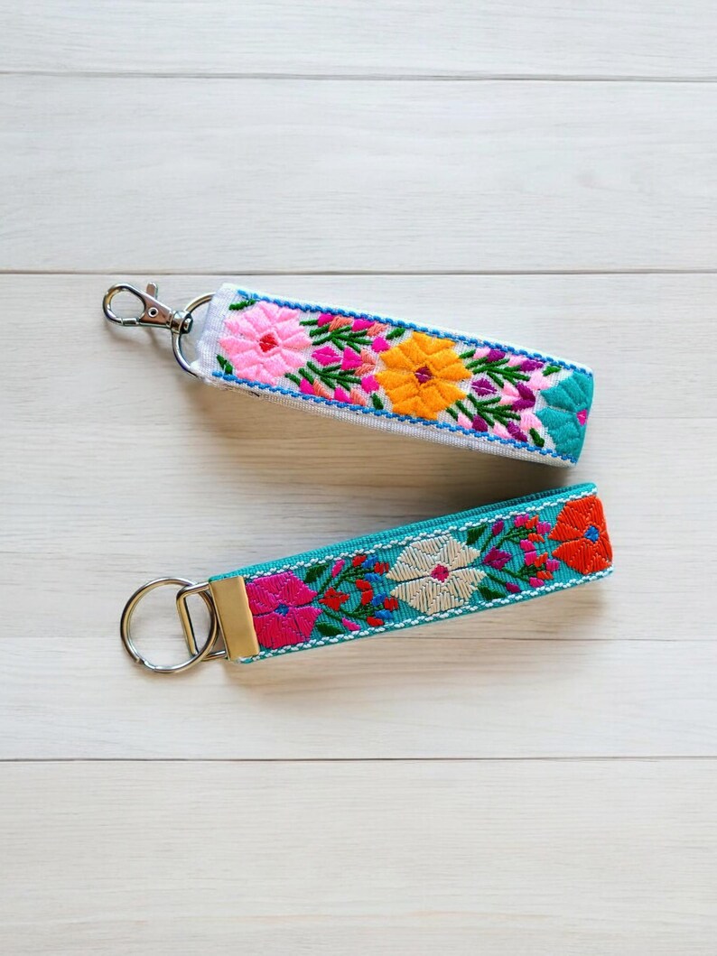 May include: Two colorful embroidered keychains with metal rings. One keychain is white with pink, orange, and yellow flowers. The other keychain is turquoise with pink, orange, and white flowers.