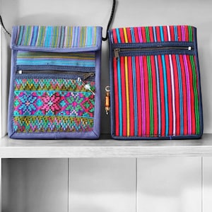 May include: Two small, rectangular crossbody bags with colorful woven patterns. One bag features vertical stripes in red, yellow, and blue. The other has a blue base with horizontal stripes and a decorative floral design. Both have zippers and adjustable straps.