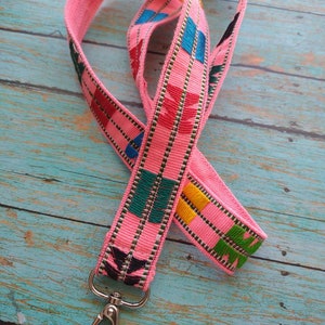 May include: A pink fabric lanyard featuring a woven geometric pattern in shades of green, blue, red, yellow, and black. The strap is constructed from a textured textile material and includes a silver-toned metal swivel lobster clasp at the end.