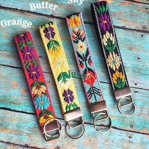 May include: Four fabric wristlet keychains with embroidered floral patterns in orange, butter yellow, sky blue, and black. Each strap features a metal clasp and a circular split ring. The text Orange, Butter, Sky, and Black labels each item.