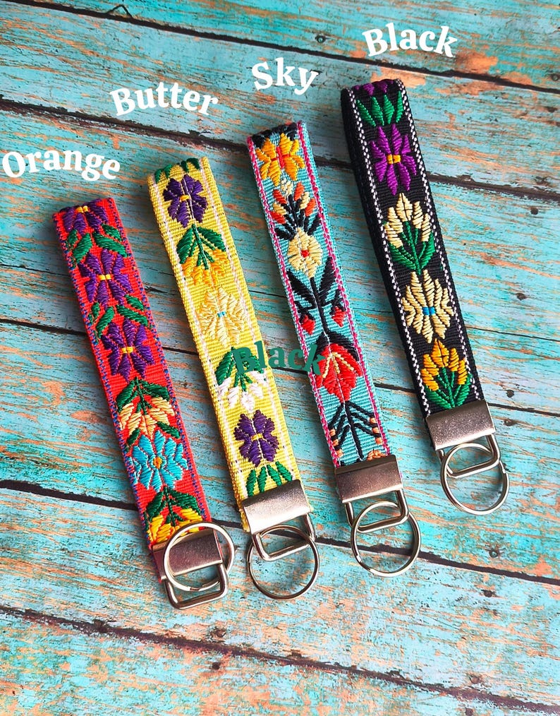 May include: Four fabric wristlet keychains with embroidered floral patterns in orange, butter yellow, sky blue, and black. Each strap features a metal clasp and a circular split ring. The text Orange, Butter, Sky, and Black labels each item.