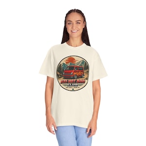 May include: An off-white t-shirt featuring a circular graphic of a red van with flame decals on a mountain road under a sunset. The text reads KEI HOT ROD CLUB and RIDE SMALL - ROAM FAR, with a small pine tree icon at the bottom of the design.