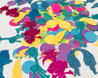 Puede incluir: Un montón de confeti de colores con siluetas de personajes de dibujos animados cubiertas de purpurina y círculos sólidos. Los personajes incluyen una jirafa, un troll y una palmera. Los colores incluyen rosa, azul, verde, amarillo y morado.