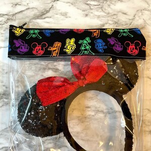May include: A clear, zippered pouch containing a black sequined headband with a red sequined bow. The top of the pouch is black fabric with colorful cartoon character faces. The pouch is designed to store and protect the headband.
