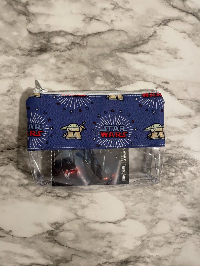 May include: A blue zippered pouch with a clear plastic front. The pouch has a repeating pattern of a cartoon character from Star Wars: The Mandalorian, the words "Star Wars", and stars. The clear front shows a Disneyland ticket for Star Wars: Galaxy's Edge.
