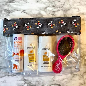 May include: Clear plastic zippered pouch with a black and white Mickey Mouse print fabric lining. The pouch contains three bottles of hair care products: Pantene Moisture Renewal Shampoo, Pantene Moisture Renewal Conditioner, and a travel-sized bottle of Sport Sunscreen Spray SPF 30. A pink Wet Brush hairbrush is also included.