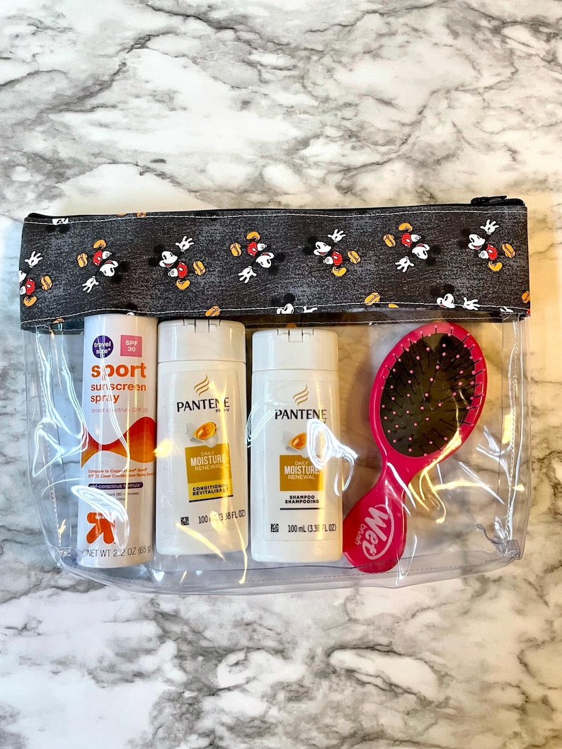May include: Clear plastic zippered pouch with a black and white Mickey Mouse print fabric lining. The pouch contains three bottles of hair care products: Pantene Moisture Renewal Shampoo, Pantene Moisture Renewal Conditioner, and a travel-sized bottle of Sport Sunscreen Spray SPF 30. A pink Wet Brush hairbrush is also included.