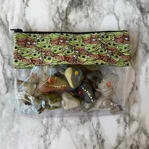 Puede incluir: Una bolsa transparente con cremallera de pl&aacute;stico transparente con una solapa de tela verde y marr&oacute;n con un patr&oacute;n repetido de un personaje de dibujos animados. La bolsa contiene varias peque&ntilde;as figuras de dinosaurios.