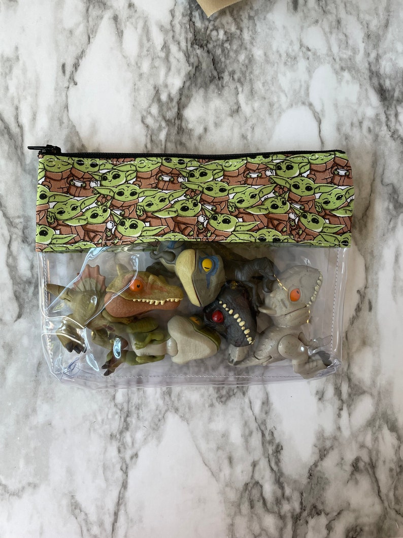 Puede incluir: Una bolsa transparente con cremallera de pl&aacute;stico transparente con una solapa de tela verde y marr&oacute;n con un patr&oacute;n repetido de un personaje de dibujos animados. La bolsa contiene varias peque&ntilde;as figuras de dinosaurios.