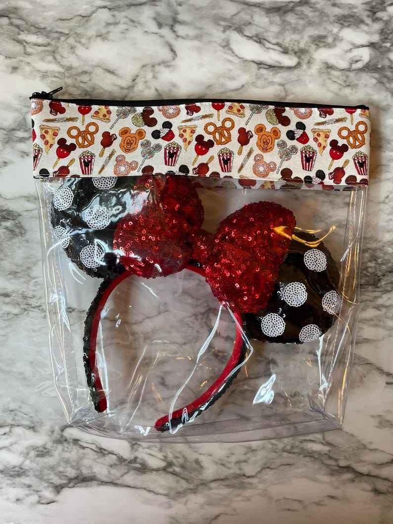 May include: A clear zipped pouch containing a pair of black and white polka dot mouse ears with a red sequined bow. The top of the pouch has a white fabric border with a food-themed pattern, including pizza, pretzels, and Mickey Mouse.
