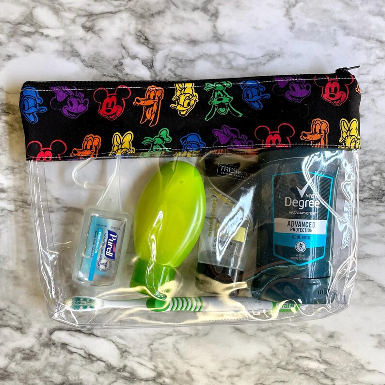 May include: A clear travel cosmetic bag with a black top featuring colorful cartoon character designs. The bag contains toiletries, including a green hairbrush, deodorant, toothpaste, and hand sanitizer.