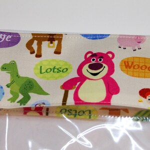May include: A clear plastic zippered pouch with a white fabric lining featuring a colorful print of characters from the Toy Story movie franchise. The print includes characters like Woody, Buzz Lightyear, Rex, and Lotso. The pouch has a zipper closure and a clear plastic front.