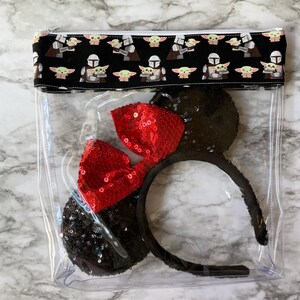 May include: Clear plastic bag with a zipper closure. The bag has a black and white fabric lining with a repeating pattern of a cartoon character. The bag contains a pair of black mouse ears with a red sequined bow.