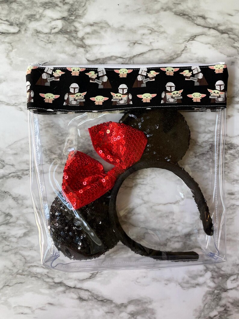 May include: Clear plastic bag with a zipper closure. The bag has a black and white fabric lining with a repeating pattern of a cartoon character. The bag contains a pair of black mouse ears with a red sequined bow.