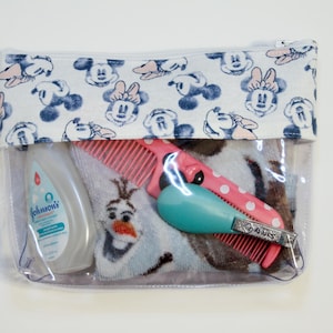 May include: Clear plastic zippered pouch with a white fabric lining featuring a repeating pattern of blue and pink Mickey Mouse heads. The pouch contains a blue and white towel, a pink comb, a blue and white bottle of Johnson's baby lotion, a pink comb, and a silver nail clipper.