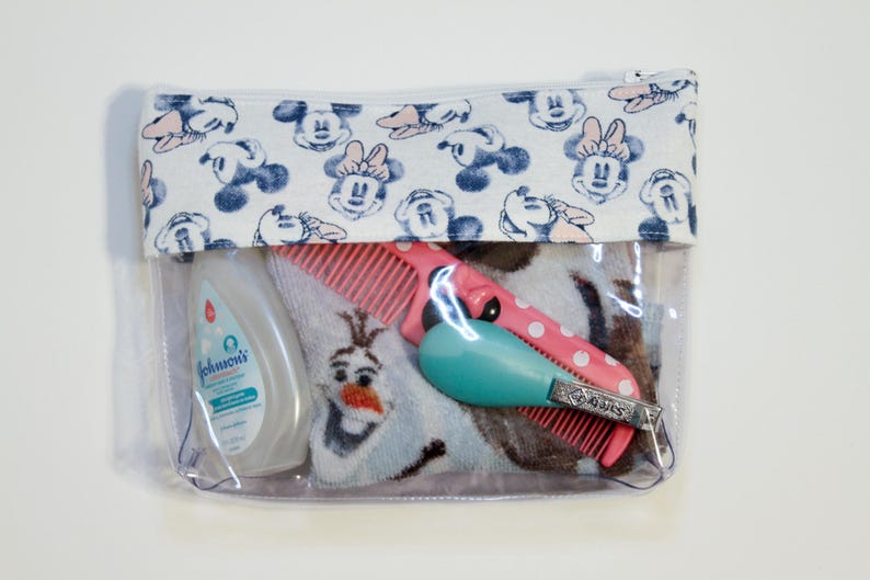 May include: Clear plastic zippered pouch with a white fabric lining featuring a repeating pattern of blue and pink Mickey Mouse heads. The pouch contains a blue and white towel, a pink comb, a blue and white bottle of Johnson's baby lotion, a pink comb, and a silver nail clipper.