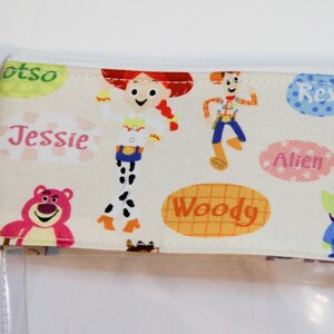 May include: A clear plastic zippered pouch with a white fabric lining featuring a Toy Story theme. The fabric has a repeating pattern of characters including Woody, Jessie, Buzz Lightyear, Rex, and the Alien. The fabric also has the names of the characters written on it.