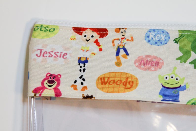 May include: A clear plastic zippered pouch with a white fabric lining featuring a Toy Story theme. The fabric has a repeating pattern of characters including Woody, Jessie, Buzz Lightyear, Rex, and the Alien. The fabric also has the names of the characters written on it.