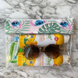 May include: A clear plastic zippered pouch with a pink and green floral print featuring a cartoon character. The pouch contains two bottles of sunscreen, a pair of sunglasses, and a small bottle of aloe vera.
