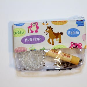 May include: A clear plastic zippered pouch with a white fabric lining featuring Toy Story characters and the words "Lotso", "Bullseye", and "Hamm". The pouch contains a clear hair scrunchie, bobby pins, and a Burt's Bees lip balm.