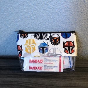 Puede incluir: Una bolsa blanca con cremallera y un frente de pl&aacute;stico transparente. La bolsa est&aacute; decorada con un patr&oacute;n repetido de cascos mandalorianos coloridos. La bolsa contiene un paquete de vendas adhesivas de la marca Band-Aid. El paquete dice "Band-Aid Brand Adhesive Bandages" y "Johnson & Johnson".