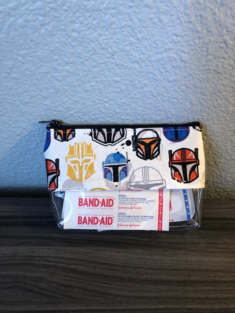 Puede incluir: Una bolsa blanca con cremallera y un frente de pl&aacute;stico transparente. La bolsa est&aacute; decorada con un patr&oacute;n repetido de cascos mandalorianos coloridos. La bolsa contiene un paquete de vendas adhesivas de la marca Band-Aid. El paquete dice "Band-Aid Brand Adhesive Bandages" y "Johnson & Johnson".