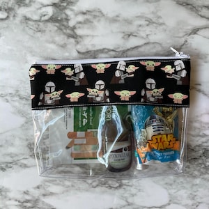 May include: A clear plastic zipped pouch with a black fabric lining featuring a repeating pattern of a Mandalorian and a Baby Yoda. The pouch contains a small bottle of hand sanitiser, a packet of tissues, and a packet of plasters.