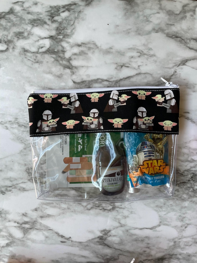 May include: A clear plastic zipped pouch with a black fabric lining featuring a repeating pattern of a Mandalorian and a Baby Yoda. The pouch contains a small bottle of hand sanitiser, a packet of tissues, and a packet of plasters.