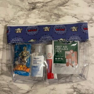 May include: A clear plastic zippered pouch with a blue and white Star Wars themed fabric lining. The pouch contains a small package of Star Wars themed tissues, a bottle of equate moisturizing hand sanitizer, a tube of Hydrocortisone cream, and a small first aid kit with the text "CORAD First Aid Portable Pack".