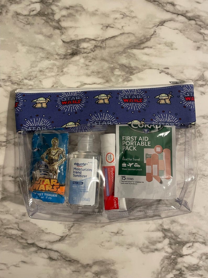 May include: A clear plastic zippered pouch with a blue and white Star Wars themed fabric lining. The pouch contains a small package of Star Wars themed tissues, a bottle of equate moisturizing hand sanitizer, a tube of Hydrocortisone cream, and a small first aid kit with the text "CORAD First Aid Portable Pack".