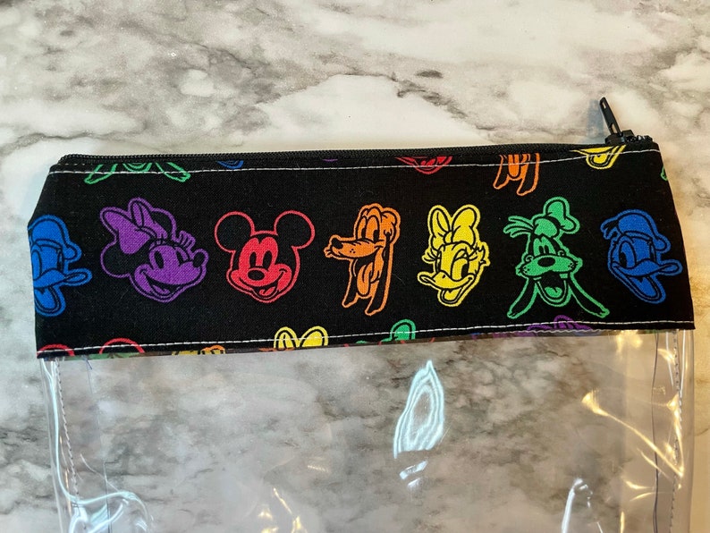 May include: A clear pencil case with a black top featuring colorful cartoon character faces. The characters include Mickey Mouse, Minnie Mouse, Donald Duck, and Goofy. The pencil case has a black zipper.