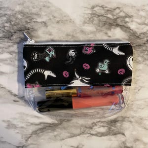 May include: A clear zippered pouch with a black fabric lining featuring a repeating pattern of characters from the Nightmare Before Christmas. The pouch is filled with two lip glosses.