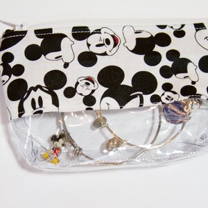 K&ouml;nnte beinhalten: Eine transparente Rei&szlig;verschlusstasche mit einem schwarz-wei&szlig;en Mickey-Mouse-Muster auf dem oberen Teil. Die Tasche enth&auml;lt mehrere silberne Armb&auml;nder, darunter eines mit einem Mickey-Mouse-Anh&auml;nger. Der Rei&szlig;verschluss ist wei&szlig;.