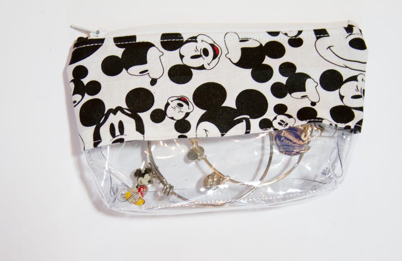K&ouml;nnte beinhalten: Eine transparente Rei&szlig;verschlusstasche mit einem schwarz-wei&szlig;en Mickey-Mouse-Muster auf dem oberen Teil. Die Tasche enth&auml;lt mehrere silberne Armb&auml;nder, darunter eines mit einem Mickey-Mouse-Anh&auml;nger. Der Rei&szlig;verschluss ist wei&szlig;.