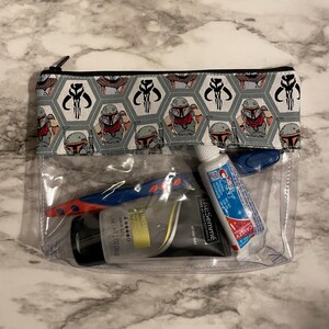 Op de afbeelding: Een doorzichtige, met een rits te sluiten tas van plastic met een grijze en zwarte, gepatroneerde voering van stof. De tas bevat een blauwe en rode tandenborstel, een tube tandpasta en een fles haar gel. De stoffen voering heeft een herhalend patroon van een cartoonfiguur met een helm en hoorns.