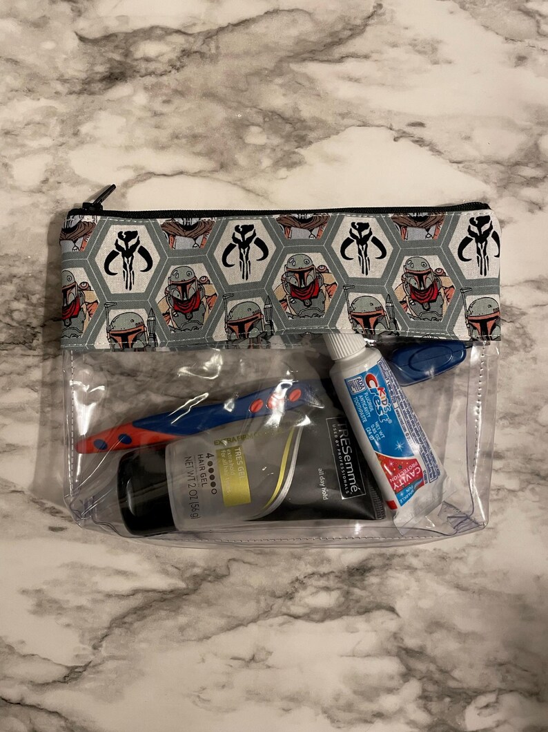 Op de afbeelding: Een doorzichtige, met een rits te sluiten tas van plastic met een grijze en zwarte, gepatroneerde voering van stof. De tas bevat een blauwe en rode tandenborstel, een tube tandpasta en een fles haar gel. De stoffen voering heeft een herhalend patroon van een cartoonfiguur met een helm en hoorns.