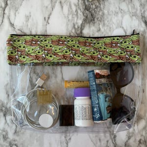 Puede incluir: Una bolsa transparente de pl&aacute;stico con cremallera, con una cremallera negra y un forro de tela verde y marr&oacute;n con un patr&oacute;n repetido de personajes de dibujos animados de Baby Yoda. La bolsa contiene un cable de carga blanco, una peque&ntilde;a botella de pastillas, un peque&ntilde;o tubo amarillo, un objeto rectangular negro y unas gafas de sol negras.